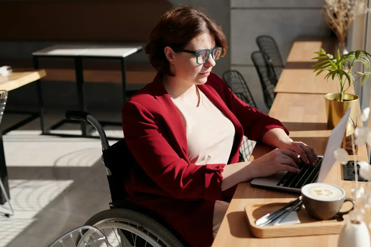  Frau mit Brille im Rollstuhl arbeitet an einem Laptop und repräsentiert den Zugang zu digitalen Inhalten für Menschen mit Behinderungen.