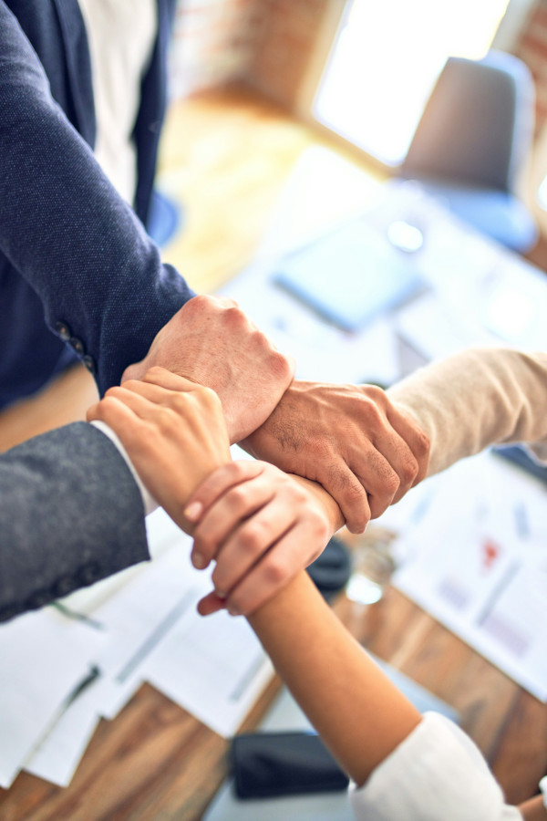 Vier Personen umgreifen gegenseitig ihre Handgelenke und bilden ein Viereck – symbolisiert Teamarbeit, Vertrauen und gegenseitige Unterstützung.